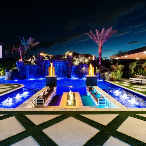 Luxury Gilbert backyard with custom grotto waterfall, blue pool lighting, fire columns, sunken fire pit, turf inlays, and outdoor lounge areas.